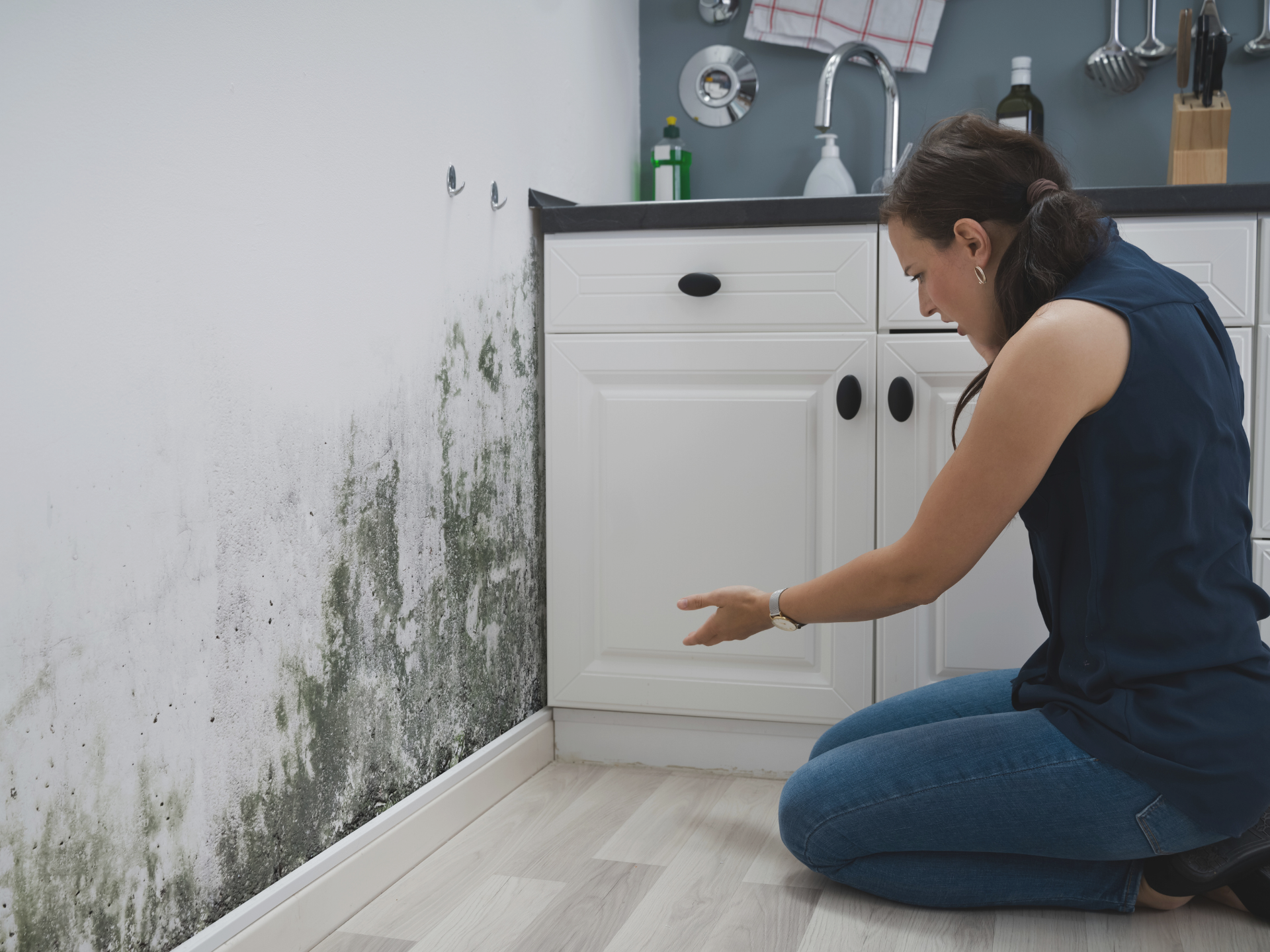 Woman kneeling in a Phoenix kitchen pointing to black mold spreading up a white wall next to the cabinet